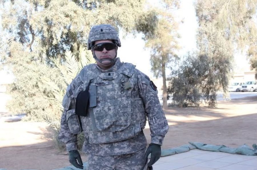 Ephraim Travis in combat gear, helmet and sunglasses, standing among palm trees in Iraq